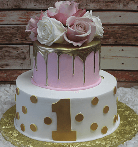 The TC-010 is a two-tier cake with a white bottom layer featuring gold polka dots and a large gold number one, a pink top layer with gold drip and pink and white roses, all set on a gold cake board.