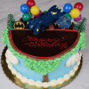 The SC-056 Batman cake is a round birthday cake with blue and green icing, topped with a Batman figure, Batmobile, balloons, trees, and "Happy Birthday" written in red on a chocolate plaque.