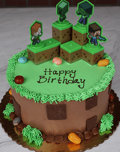 The SC-005 Minecraft cake is round with green and brown frosting, block shapes, four Minecraft character toppers, “Happy Birthday” on top, and candy decorations around the base.