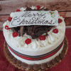 The CH-008 Display Black Forest with Christmas Decor is a round cake topped with white frosting, chocolate shavings, cherries, a plaid ribbon around the side, and a white “Merry Christmas” plaque on top.