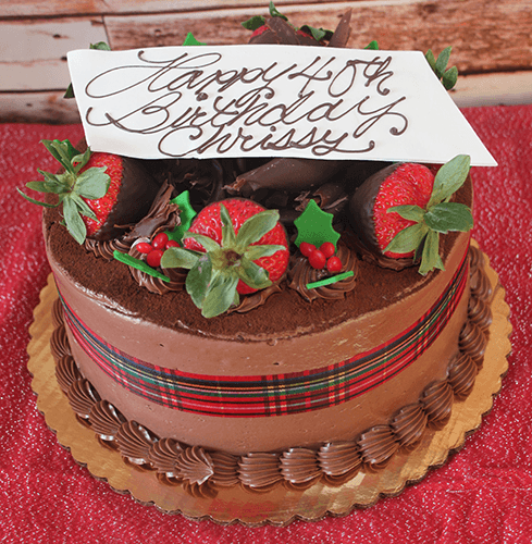 A CH-006 Display Tiramisu with Christmas decor, adorned with a plaid ribbon, chocolate-dipped strawberries, red berries, and a white chocolate plaque reading "Happy 40th Birthday Chrissy," on a gold cake board.