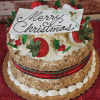 Round carrot cake with cream cheese filling, decorated with festive Christmas accents. Features a white chocolate "Merry Christmas" plaque, plaid ribbon wrap, and presented on a gold board. Product Name: CH-003.