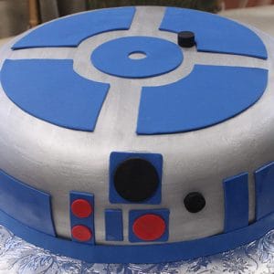The CB-007B is a round cake styled like R2-D2 from Star Wars, featuring silver-gray icing with blue, red, and black fondant accents, displayed on a silver patterned tray.
