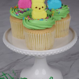A gold cupcake with chocolate filling is displayed on a white cake stand, surrounded by green decorative grass in the foreground.