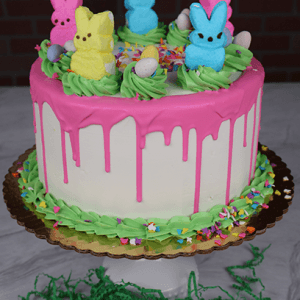 The EA-000B is a white frosted cake with pink drip icing, topped with colorful marshmallow bunny candies, mini eggs, and green frosting. Sprinkles decorate the base, and it sits on a gold board with green artificial grass.