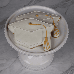 Two GC-0002 cookies, decorated as white graduation caps with yellow tassels, are displayed on a white cake stand against a marble background.