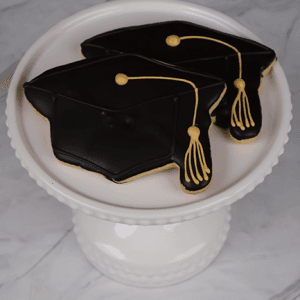 Two GC-0001 cookies, decorated as black graduation caps with yellow tassels, are displayed on a white cake stand against a marble background.