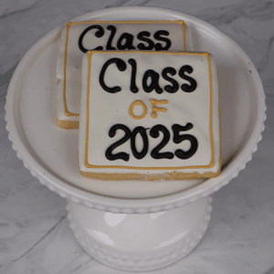 Two square cookies, GC-0003, feature white icing with black and gold "Class of 2025" lettering and are displayed on a decorative white cake stand against a marble background.