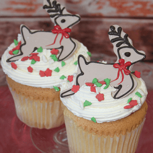Two CH-067A gold cupcakes with chocolate mousse filling, decorated with white frosting, red and green star sprinkles, and topped with festive reindeer-shaped cookies adorned with red bows.