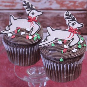 Two CH-067B Chocolate cupcakes with white chocolate mousse filling, decorated with star sprinkles and reindeer-shaped cookies with red bows, displayed on a glass stand.