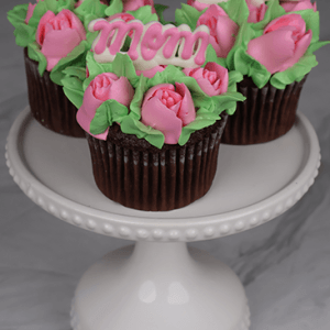 Three MD-000B chocolate cupcakes with white mousse filling, topped with pink mom toppers and rose-shaped frosting, displayed on a white beaded-edge cake stand against a marble background.