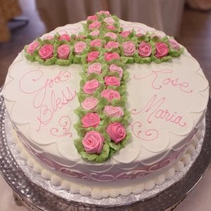 RC-032 is a round white cake featuring a cross of pink and green icing roses. "God Bless" and "Rose Maria" are written in pink icing, with the edges finished in white piped details.