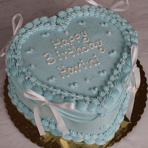 The BD-023G Heart shape one size cake features blue icing, white decorations, small hearts, pearls, bows, and "Happy Birthday Harini" on top. It is presented on a gold board.