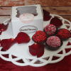A white box labeled “VC-016 Chocolate Bailey Truffles” rests on a tray with four decorated truffles and scattered red rose petals, set against a rustic wooden background.