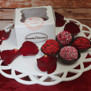 A white box labeled “VC-016 Chocolate Bailey Truffles” rests on a tray with four decorated truffles and scattered red rose petals, set against a rustic wooden background.