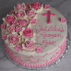 RC-018 is a round cake with pink and white icing roses, a pink cross, "God Bless Reagan Marie" in pink frosting, plus pink swirls and small flowers along the bottom edge.