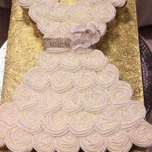 The BR-100 features cupcakes arranged and decorated with white icing to resemble a wedding dress, accented by a flower and jeweled belt detail, and displayed on a gold-patterned board.