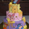 The BB-000A is a three-tiered cake featuring Winnie the Pooh, Piglet, Tigger, and Eeyore, with honeycomb accents, dripping honey, buzzing bees, and a tilted "Hunny" pot topper.