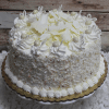 The BD-018H Marble Coconut is a round, layered cake with white frosting, shredded coconut, decorative swirls, white chocolate curls, and small white candies, beautifully presented on a cake stand against a rustic wood background.