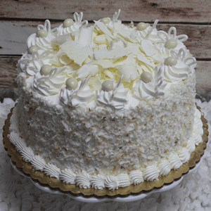 The BD-018H Marble Coconut is a round, layered cake with white frosting, shredded coconut, decorative swirls, white chocolate curls, and small white candies, beautifully presented on a cake stand against a rustic wood background.
