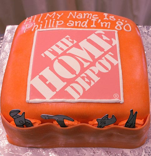 CO-009: A square orange cake with The Home Depot logo centered, icing tools on the sides, and "Hi! My name is Phillip and I'm 80." written on top, displayed on a silver surface.