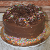 The BD-018J Gold cake with chocolate mousse filling features a round shape, swirled chocolate frosting, chocolate shavings, and confetti sprinkles on a gold cake board against a rustic wooden background.
