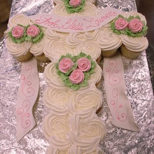 The RC-110 is a cross-shaped cake made of white frosted cupcakes, decorated with pink and green flowers and a white ribbon with “God Bless Lauren” in pink icing, displayed on a silver foil-covered tray.