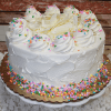 The BD-018K Gold cake features a round white frosted design with whipped cream filling, topped with swirls, white chocolate shavings, and colorful sprinkles on a gold cake board and white stand.