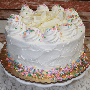 The BD-018K Gold cake features a round white frosted design with whipped cream filling, topped with swirls, white chocolate shavings, and colorful sprinkles on a gold cake board and white stand.