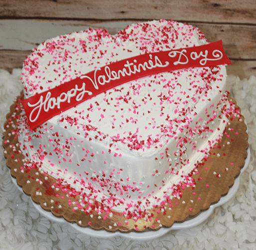 The VC-012 is a heart-shaped cake topped with white icing, pink and red sprinkles, and a red "Happy Valentines Day" banner in white script, presented on a gold cake board.