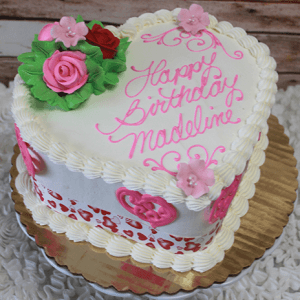 The VC-013A is a heart-shaped cake featuring pink and red flowers, green leaves, white icing borders, pink heart designs, and "Happy Birthday Madeline" written in pink icing.