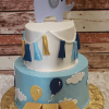TC-012 is a two-tier cake featuring an elephant with a "1" balloon topper, tassels, clouds, and side balloons. Yellow blocks spelling “JAY” decorate the base. Displayed on a gold board against a rustic wood background.