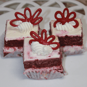 Three AP-045G Red Velvet Squares with white frosting, whipped cream, and red chocolate flower decorations are arranged on a white plate.