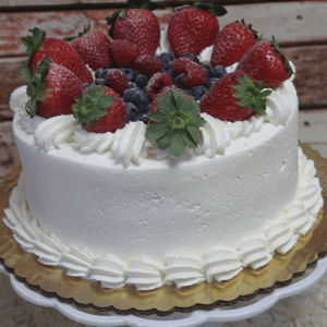 The BD-018M Tres Leches is a round cake covered in white frosting, adorned with whipped cream swirls and topped with fresh strawberries, blueberries, and raspberries, displayed on a gold-rimmed cake board.
