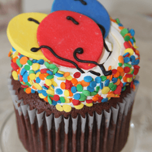 The CC-121 chocolate cupcake with white filling is topped with white frosting, rainbow sprinkles, and three fondant balloons in red, yellow, and blue with black icing strings, served on a glass plate.