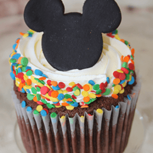The CC-125 chocolate cupcake with white filling features white frosting, colorful round sprinkles, and a Mickey Mouse ear-shaped black cookie on top, displayed on a glass plate.