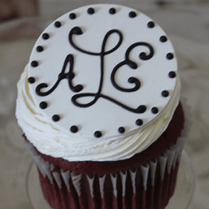 A CC-133 chocolate cupcake with white filling, topped with a round fondant disc featuring black dots and the initials ALE in black icing.