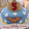 A CB-006H Wonder Woman cake with blue icing, white stars, a yellow and red logo, a yellow crown featuring a red number 5 on top, and a "Happy Birthday Zuleily" sign.