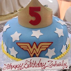 A CB-006H Wonder Woman cake with blue icing, white stars, a yellow and red logo, a yellow crown featuring a red number 5 on top, and a "Happy Birthday Zuleily" sign.