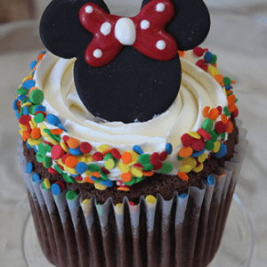 A CC-129 chocolate cupcake with white filling, topped with white frosting, colorful confetti sprinkles, and a black fondant Minnie Mouse-shaped decoration with a red bow.