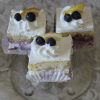 Three AP-045J Lemon Blueberry Squares—layered cake slices with white frosting, topped with whipped cream, two blueberries, and a lemon wedge—are arranged on a glass plate.