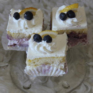 Three AP-045J Lemon Blueberry Squares—layered cake slices with white frosting, topped with whipped cream, two blueberries, and a lemon wedge—are arranged on a glass plate.