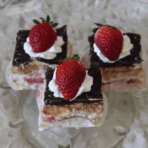 The Pina Colada Square is a three-layered dessert square topped with chocolate glaze, whipped cream, and a fresh strawberry, served on a glass plate.