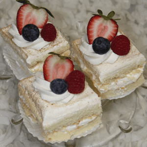 AP-045K Tres leches squares: Three sponge cake squares topped with whipped cream, strawberries, blueberries, and raspberries, served on a glass plate against a white ruffled background.