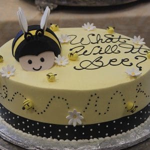 The BB-058 is a round yellow cake featuring a smiling bee face, fondant bees, white flowers, "What will it bee?" in black icing, and a black ribbon with white dots around the base.