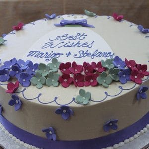 The BR-026 is a round cake with purple, red, and green flower-shaped fondant, a purple ribbon at the base, and "Best Wishes Marigo & Stefanos" written in blue icing on top.