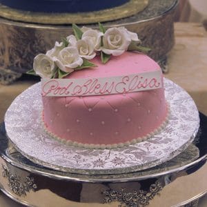 The RC-065 is a small pink cake with white icing roses and piped pearl dots, topped with “God Bless Elisa” in pink icing. Presented on a silver tray with a lacy doily underneath.