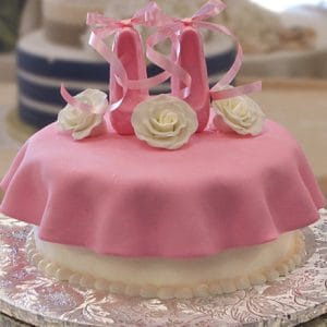 BB-060 is a round cake with pink fondant, topped with white fondant roses and pink ballet slippers. Presented on a silver board with a scalloped white base border.