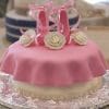 The CB-026C is a round cake covered in pink fondant with ruffled edges, topped with two pink fondant ballet slippers and three white fondant roses, all displayed on a decorative silver platter.