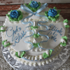 The RC-030 is a round white cake with blue and green icing roses, piped leaves, and a blue ribbon. “God Bless Elijah Cole” is written in blue script. The cake sits on a silver floral-patterned board.